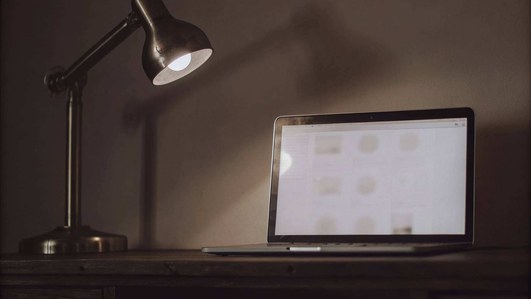 A laptop glows with a soft, blurred chat interface on a dark wood desk, lit by a warm brass desk lamp, with a manufacturing shop floor faintly visible in the background.