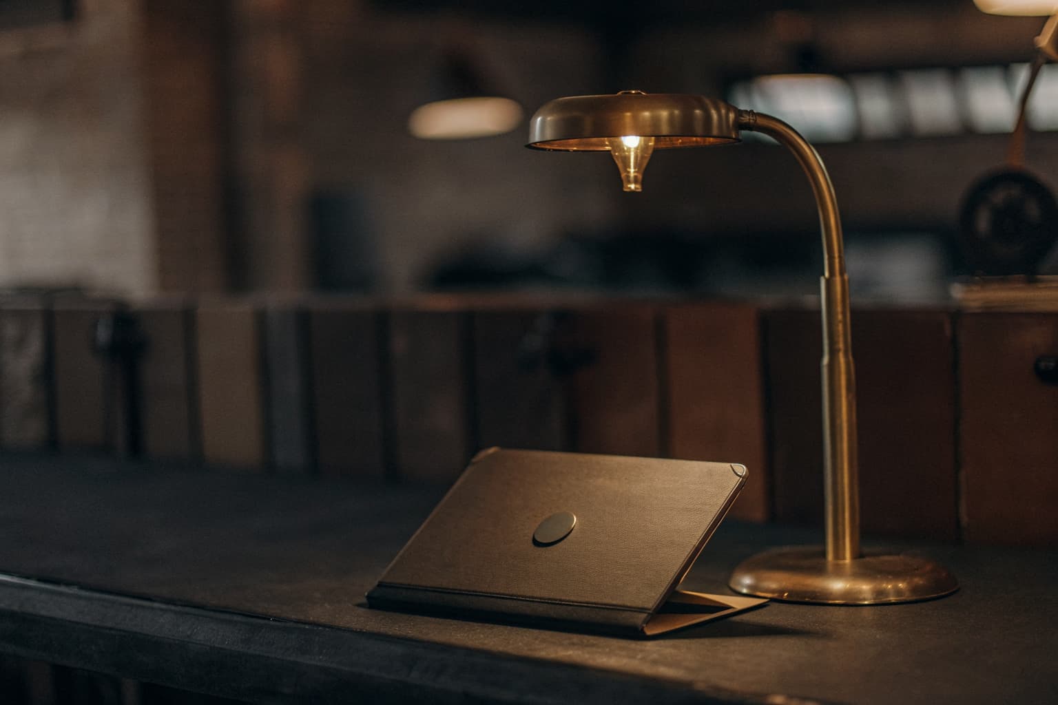 A desk lamp casts warm gold light over a closed laptop on a dark workbench, with blurred factory equipment in the background.